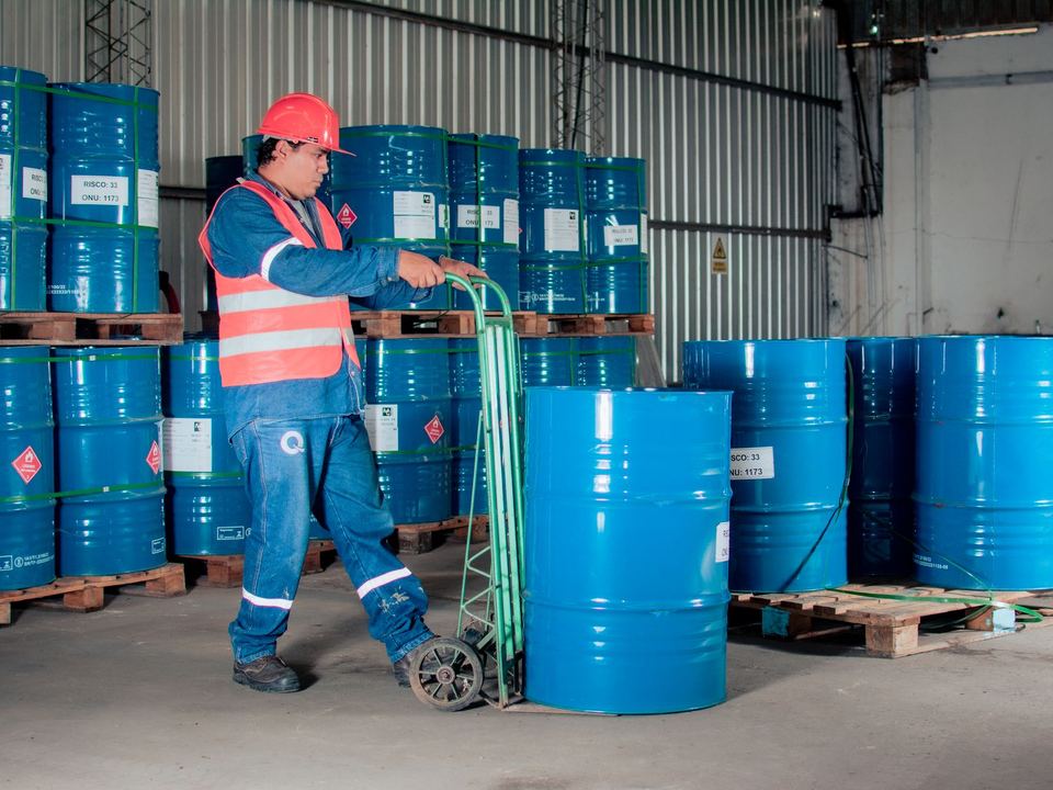 Industrial worker with a hand truck moving blue barrels in a warehouse setting.